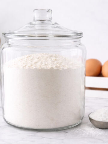 A large glass jar filled with flour sits on a marble countertop, with a metal scoop of flour beside it and a carton of brown eggs in the background.