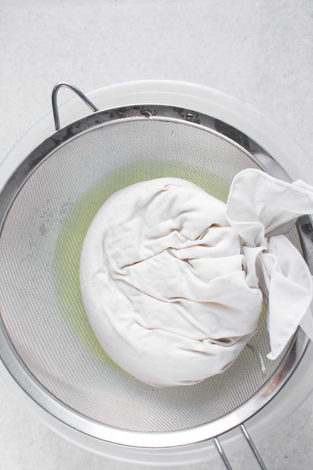 A white cloth bundle containing homemade yogurt, sitting in a metal strainer over a bowl with whey beneath the strainer.