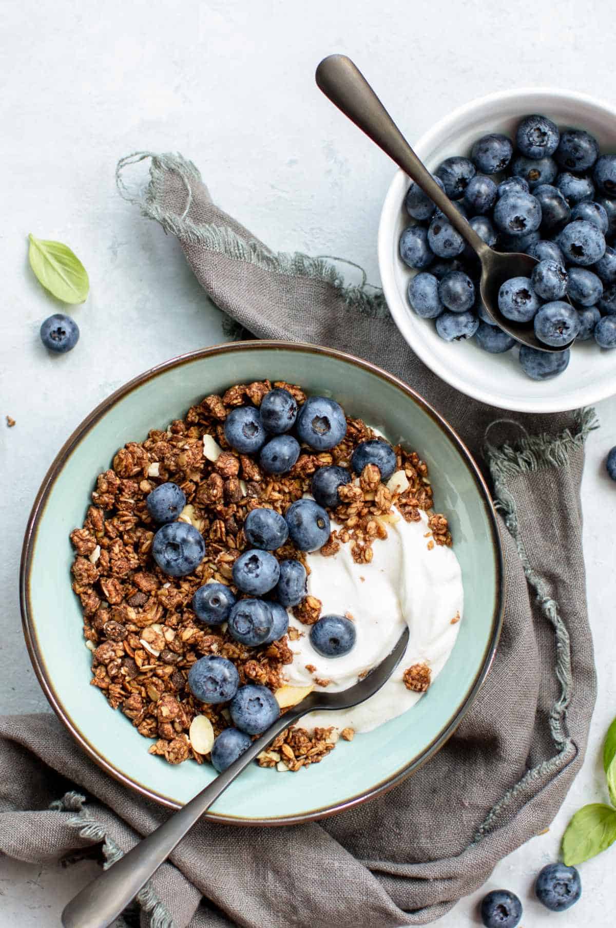 Granola, yogurt, and blueberries in a bowl with spoon on grey napkin; extra blueberries, spoons, and basil scattered nearby.