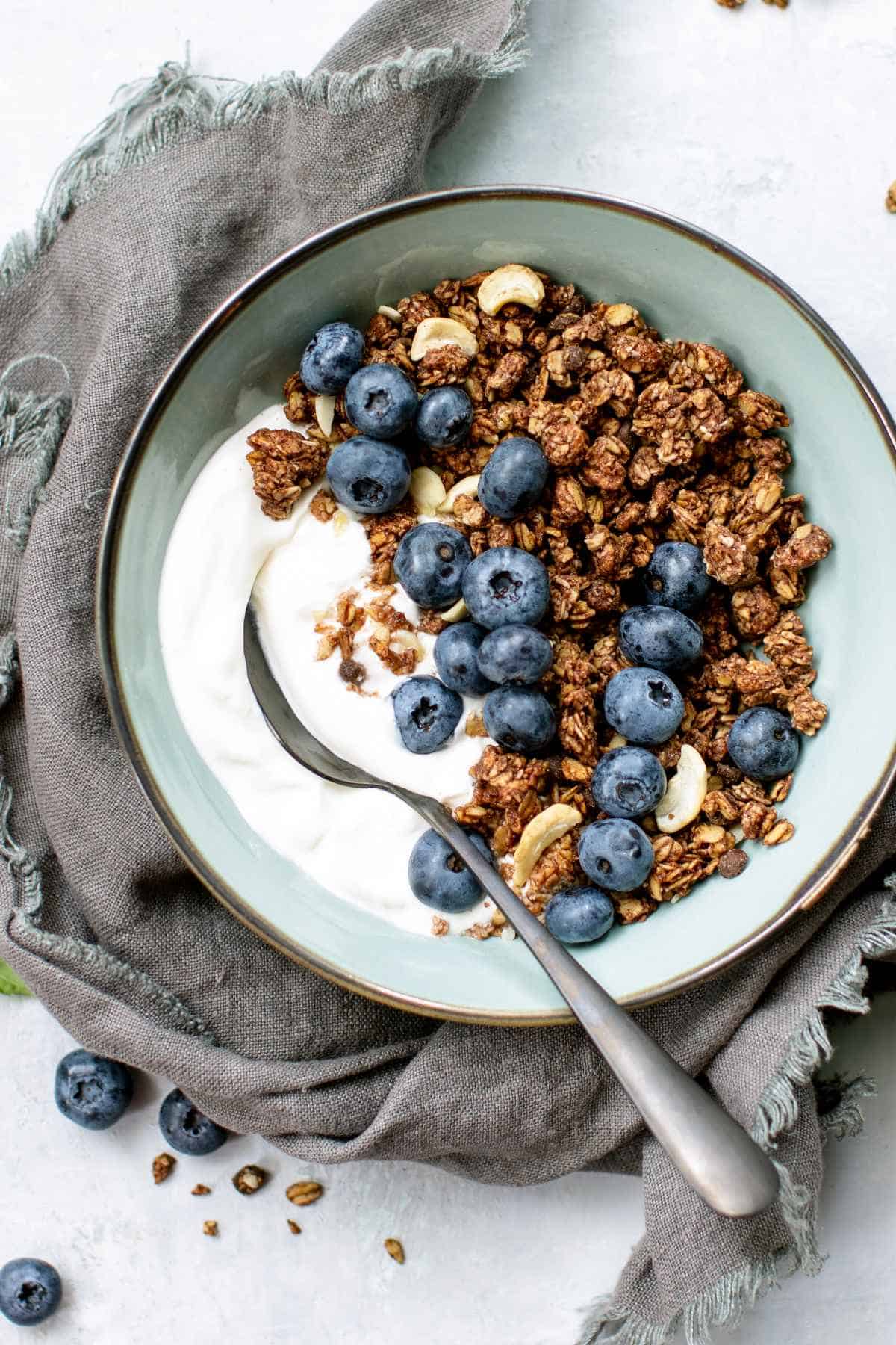 Two glass cups filled with yogurt, granola, banana slices, and nuts. A hand is drizzling honey from a spoon over the top.