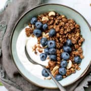 Two glass cups filled with yogurt, granola, banana slices, and nuts. A hand is drizzling honey from a spoon over the top.