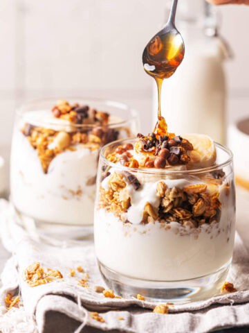 Two glass cups filled with yogurt, granola, banana slices, and nuts. A hand is drizzling honey from a spoon over the top.