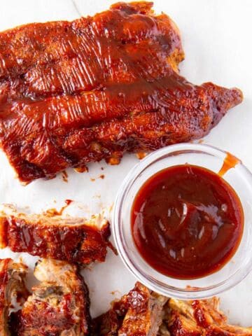 Barbecued pork ribs with a shiny glaze sit on a white surface next to a small glass bowl filled with dark red barbecue sauce. Some ribs are cut into individual pieces.