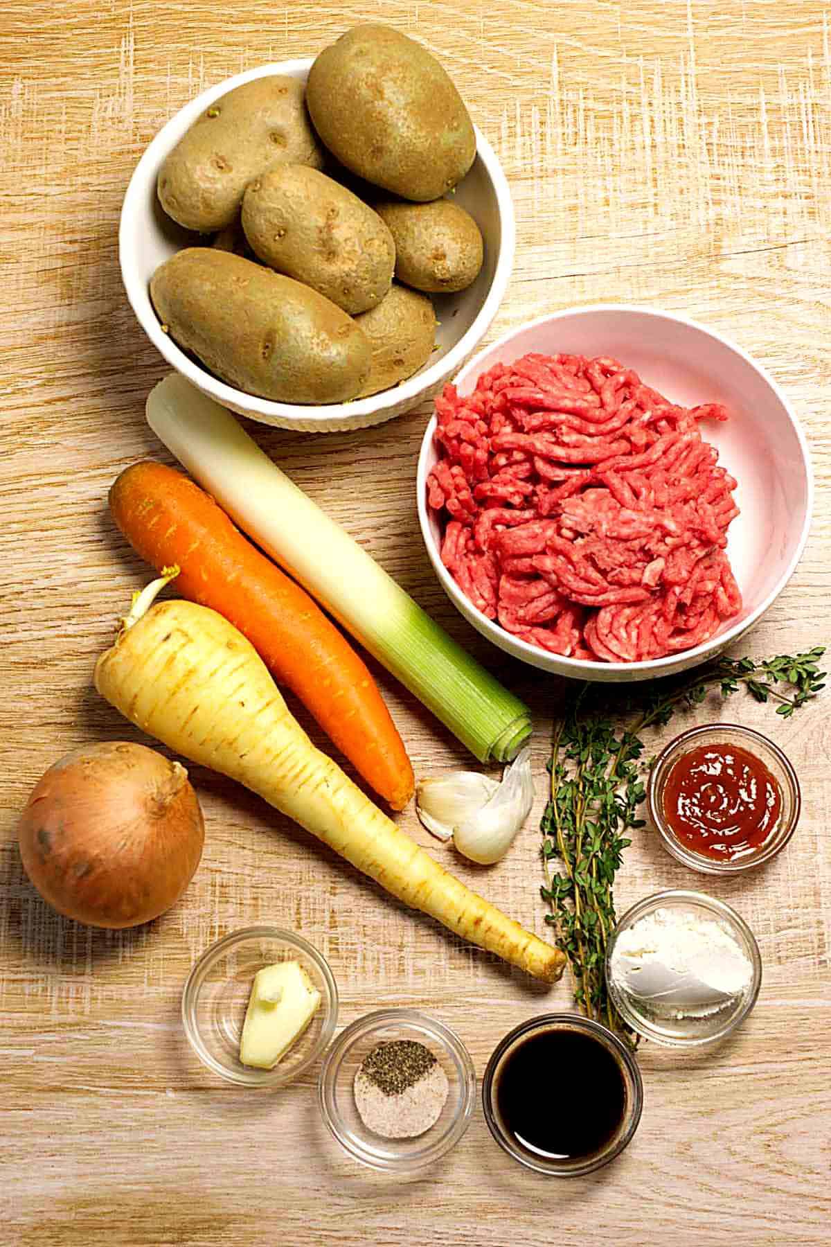 A flat lay of ingredients on a wooden surface, including potatoes, ground lamb, a leek, carrot, parsnip, onion, garlic, thyme, ketchup, cream, soy sauce, salt, and pepper in small glass bowls.