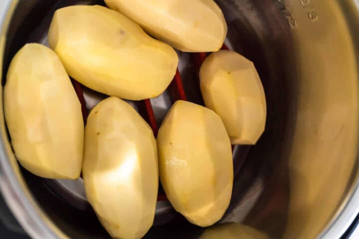 Six peeled, whole potatoes are arranged on a metal trivet inside a stainless steel pot, ready to be cooked.