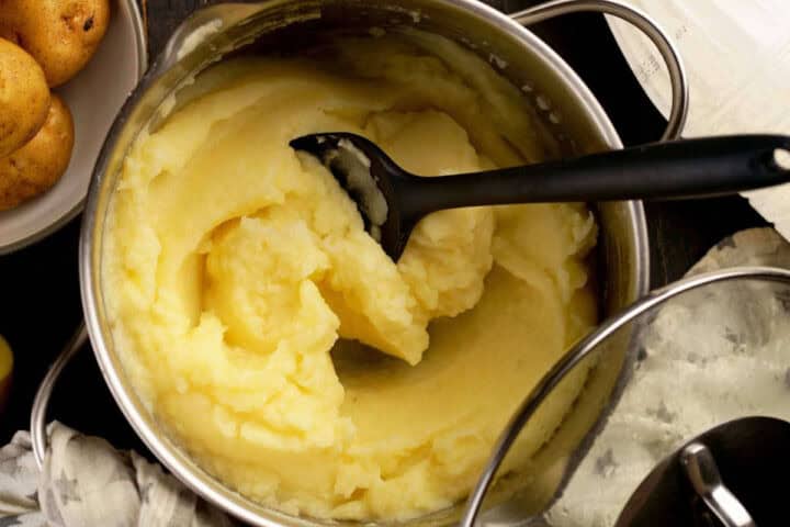 A pot of creamy mashed potatoes with a black serving spoon inside, next to a plate of whole potatoes and a glass pot lid on a dark surface.
