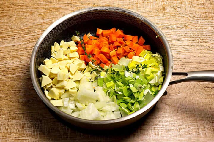 A frying pan on a wooden surface filled with chopped vegetables in sections: parsnip, carrot, leek, and onion, with a few sprigs of fresh thyme on top.