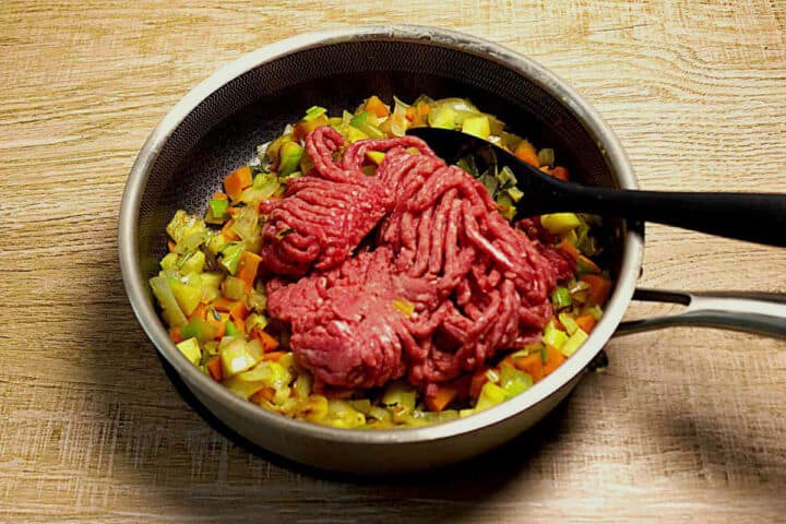A frying pan with chopped carrot, parsnip, onion, leeks and raw ground lamb, being stirred with a black spoon, on a wooden surface.