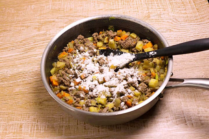 A saucepan filled with cooked ground lamb, diced carrots, parsnips, and leeks is on a wooden surface. A black spatula rests in the mixture, which is topped with a layer of white flour.