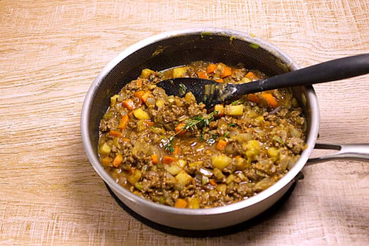 A saucepan filled with a cooked mixture of ground lamb, diced parsnips, carrots, and herbs sits on a light wooden surface. A black spoon rests in the pan, ready for serving.