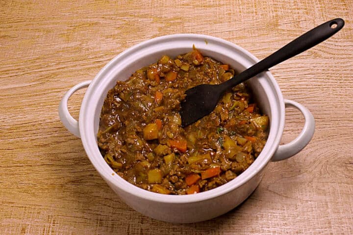 A white pot filled with a ground lamb filling containing parsnips, carrots, and diced vegetables sits on a wooden surface. A black spatula rests inside the pot.