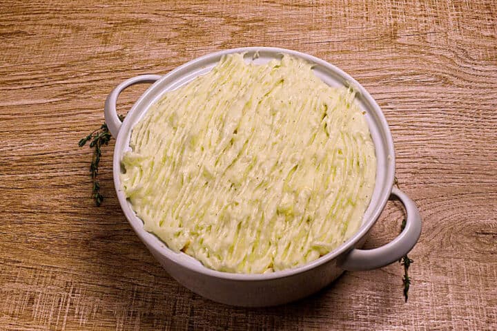 A round baking dish filled with mashed potatoes, smoothed and textured on top, sits on a wooden surface. Two sprigs of fresh herbs rest beside the handles of the dish.