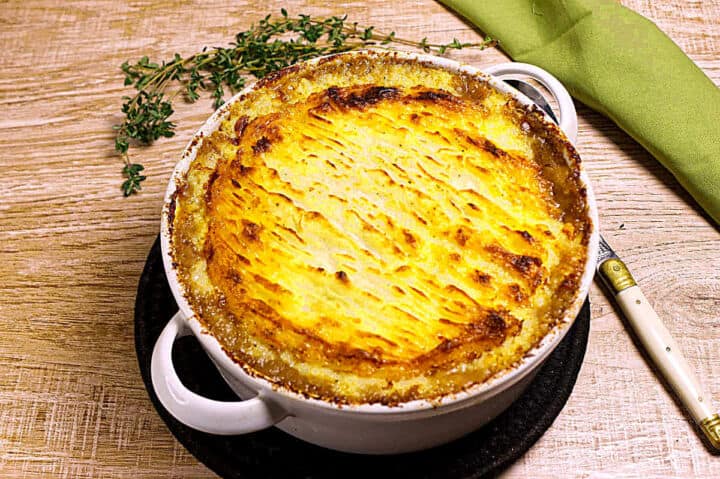 A golden-brown baked shepherd’s pie in a white casserole dish sits on a wooden table, garnished with fresh herbs and accompanied by a green napkin and a knife.