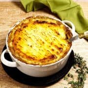 A baked shepherd's pie with a golden-brown mashed potato topping sits in a white casserole dish on a wooden table, with a serving spoon and fresh thyme nearby. A green cloth is in the background.
