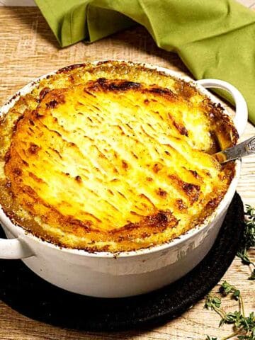 A baked shepherd's pie with a golden-brown mashed potato topping sits in a white casserole dish on a wooden table, with a serving spoon and fresh thyme nearby. A green cloth is in the background.