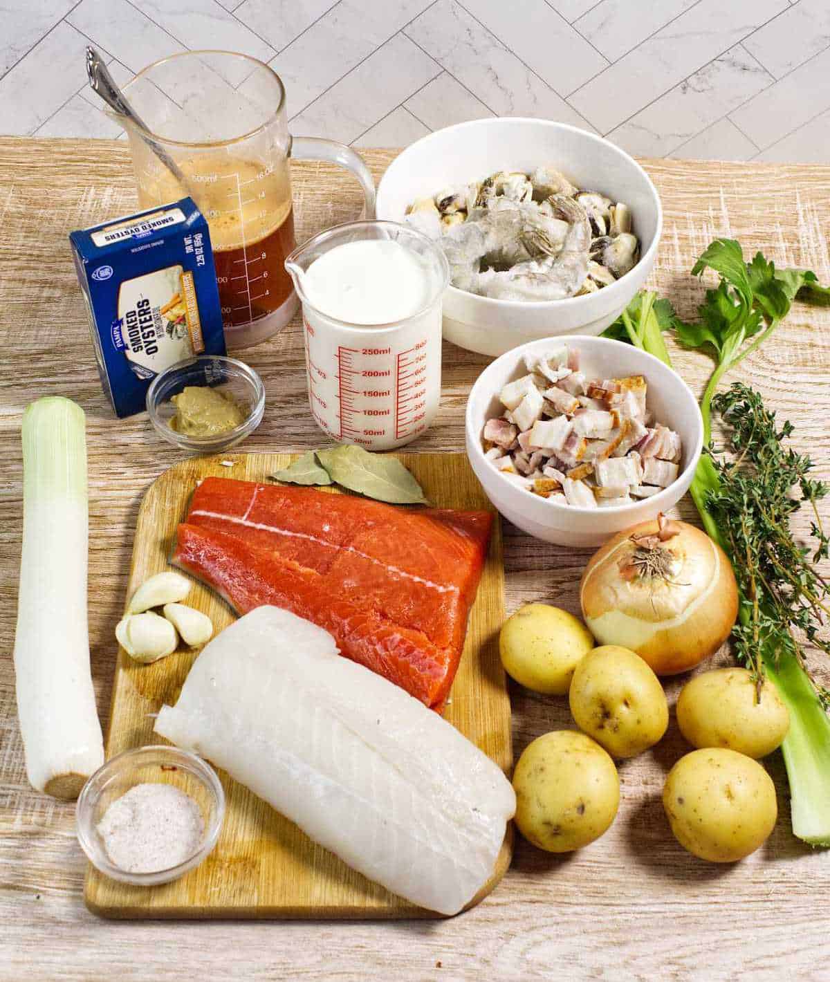 A wooden table with ingredients for fish chowder: salmon, white fish, leeks, potatoes, onion, celery, garlic, bacon, cream, seafood broth, butter, salt, bay leaves, and fresh herbs.
