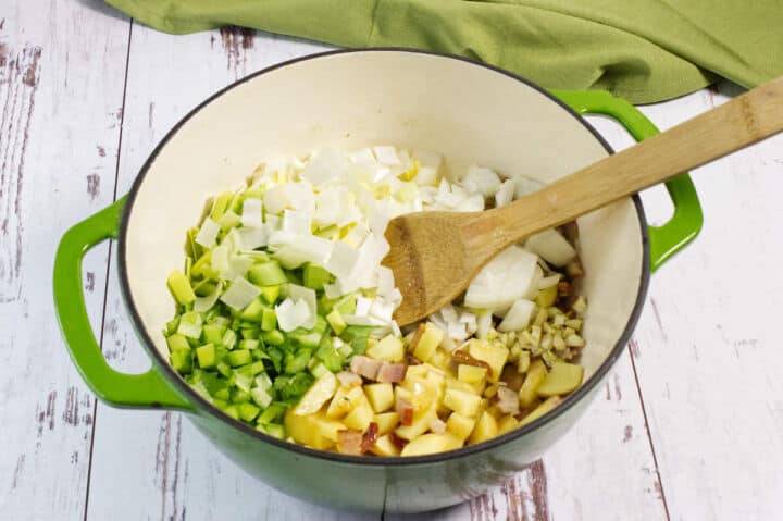 A green pot on a white wooden surface contains chopped onions, celery, zucchini, potatoes, bacon, and garlic, with a wooden spatula resting inside. A green cloth is in the background.
