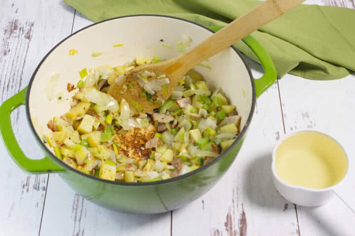 A green pot on a white wooden surface contains chopped onions, potatoes, and leeks being sautéed with a wooden spoon. A green cloth and a small white cup of cream are nearby.