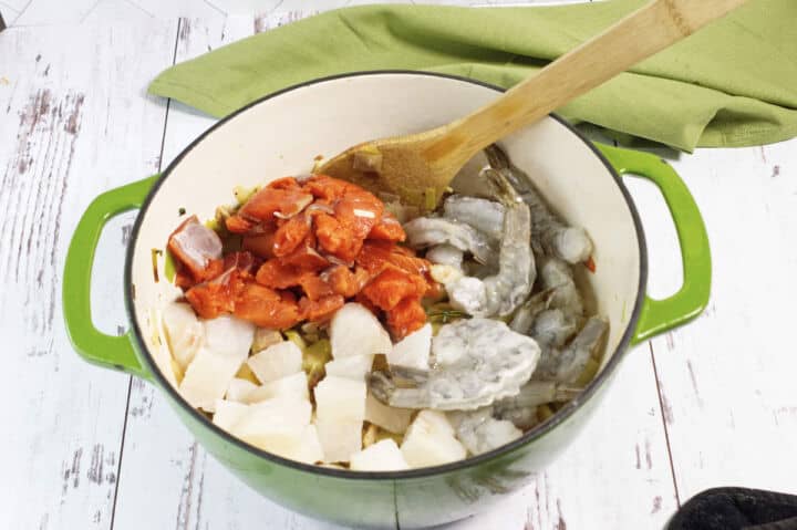 A green pot on a white wooden surface contains raw shrimp, chopped white fish, and chunks of salmon, with a wooden spoon resting inside. A green cloth is in the background.