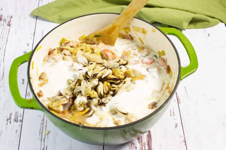 A green pot filled with seafood chowder, including mussels, cream, vegetables, and a wooden spoon, sits on a rustic white wooden surface with a green cloth in the background.