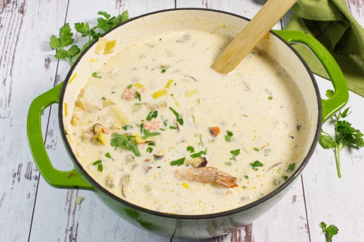 A green pot filled with creamy seafood chowder, featuring shrimp, diced potatoes, herbs, and visible parsley garnish. A wooden spoon rests inside the pot on a rustic white wooden surface.