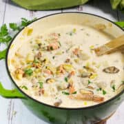 A pot of creamy seafood chowder with chunks of fish, shellfish, potatoes, and herbs sits on a white wooden surface. A wooden spoon rests in the pot, and fresh parsley is visible nearby.