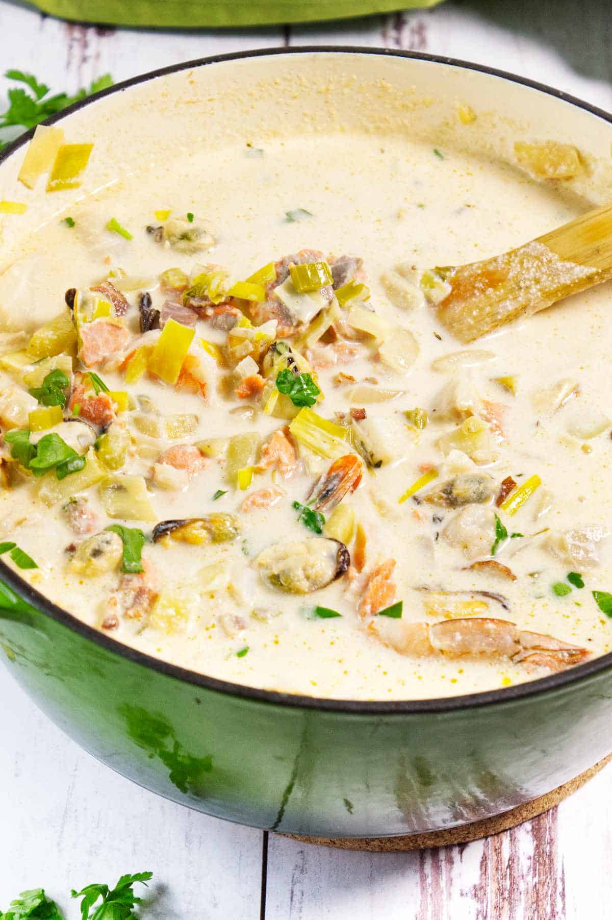 A green pot filled with creamy seafood chowder containing shrimp, clams, mussels, chopped vegetables, and herbs, with a wooden spoon stirring the mixture.
