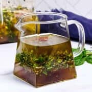 A clear glass pitcher filled with a herb-infused marinade, with visible chopped herbs and spices settled at the bottom, sits on a white surface next to fresh mint leaves.