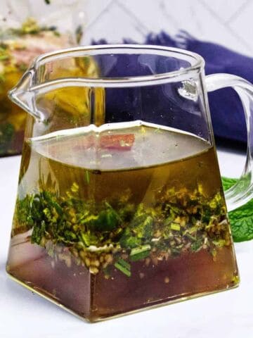 A clear glass pitcher filled with a herb-infused marinade, with visible chopped herbs and spices settled at the bottom, sits on a white surface next to fresh mint leaves.