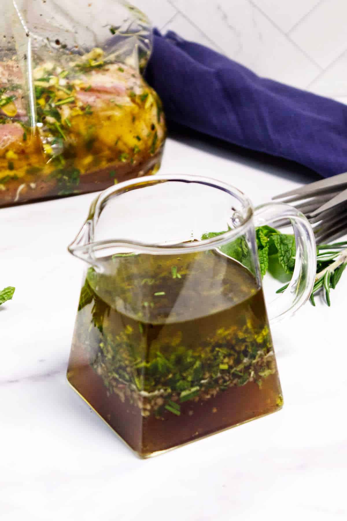 Mediterranean lamb marinade in pitcher with herbs and marinating lamb in background.