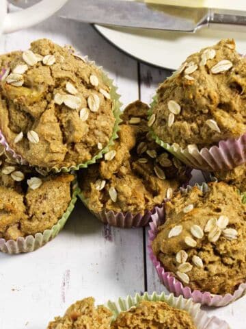 A stack of homemade muffins in colorful paper liners, topped with oats, displayed on a white wooden surface. A butter knife and plate are visible in the background.