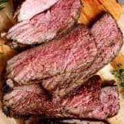 Sliced oven roasted tri-tip steak on a wooden board, showing a juicy pink center and a brown, seasoned crust. Fresh herbs are placed nearby as garnish. Text overlay reads "Oven Roasted Tri Tip.