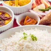 A plate of cooked basmati rice garnished with cilantro is served with bowls of Indian curry, pickles, sliced red onions, lemon, and a bread roll on a red tray. Text reads "Rice Cooker Basmati Rice.