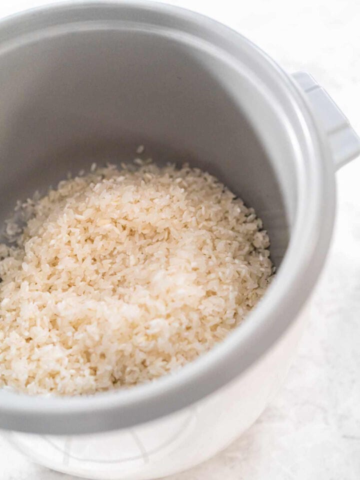 Uncooked white basmati rice in the inner pot of a rice cooker, placed on a light-colored surface.