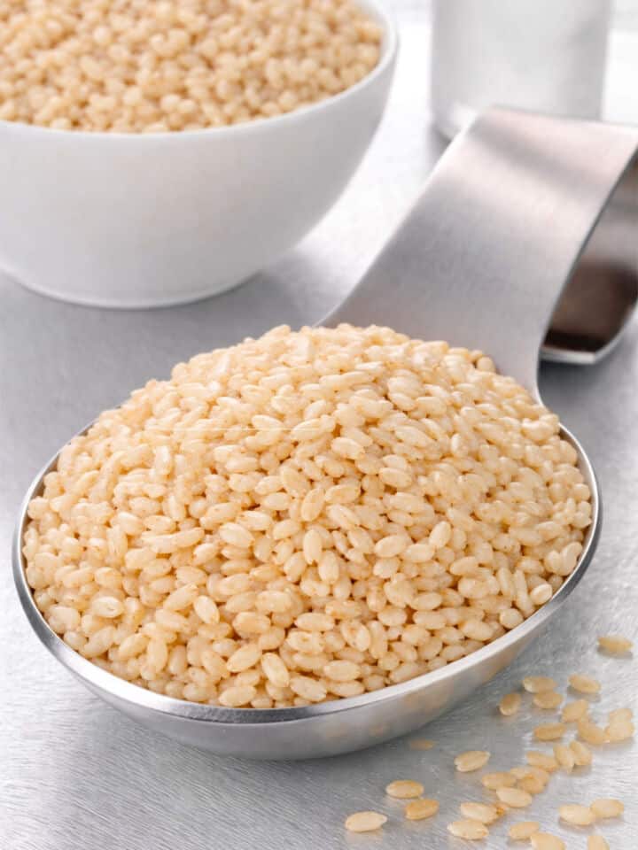 A metal scoop filled with uncooked brown rice grains sits on a light surface, with a white bowl of more rice blurred in the background.