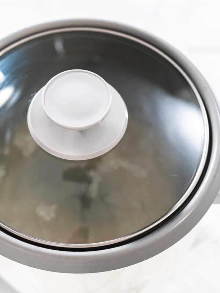 A close-up view of a pot with a glass lid, showing light reflections on the surface. The pot is placed on a light-colored countertop.
