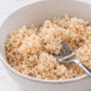 A close-up of a bowl filled with cooked brown rice, with a fork scooping some of the rice. The bowl is white with subtle speckles, placed on a light surface.