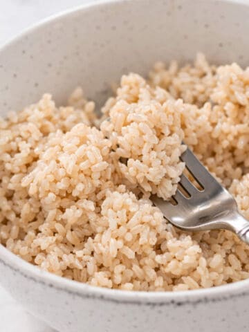 A close-up of a bowl filled with cooked brown rice, with a fork scooping some of the rice. The bowl is white with subtle speckles, placed on a light surface.