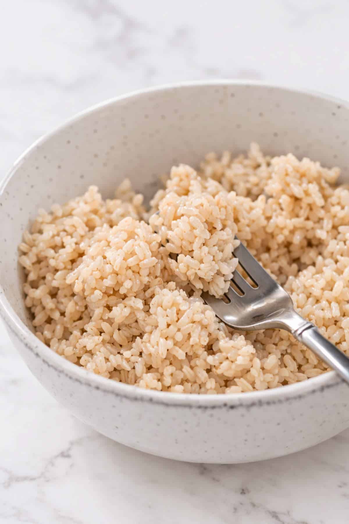 A white speckled bowl filled with cooked brown rice, with a silver fork resting inside the rice. The bowl is placed on a light-colored, marble surface.