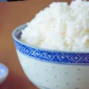 A blue and white patterned bowl filled with cooked jasmine rice sits on a wooden surface. Text above reads “Rice Cooker Jasmine Rice." Part of a website URL is visible at the bottom.