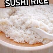 A wooden bowl filled with freshly cooked sushi rice being fluffed with a white spatula. Bold text at the top reads "Rice Cooker Sushi Rice.