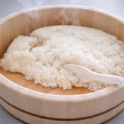 Steaming hot white rice in a round wooden tub, with a white plastic rice paddle resting on the rice.