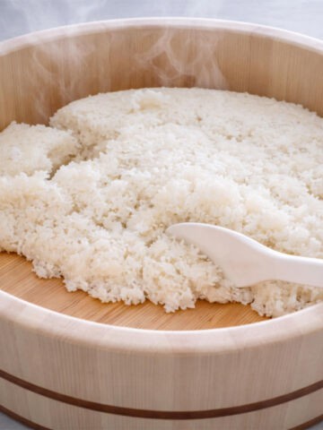 Steaming hot white rice in a round wooden tub, with a white plastic rice paddle resting on the rice.