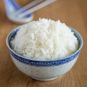 A bowl of steamed white rice sits on a wooden table, with chopsticks and a ceramic spoon blurred in the background.