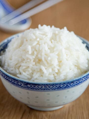 A bowl of steamed white rice sits on a wooden table, with chopsticks and a ceramic spoon blurred in the background.