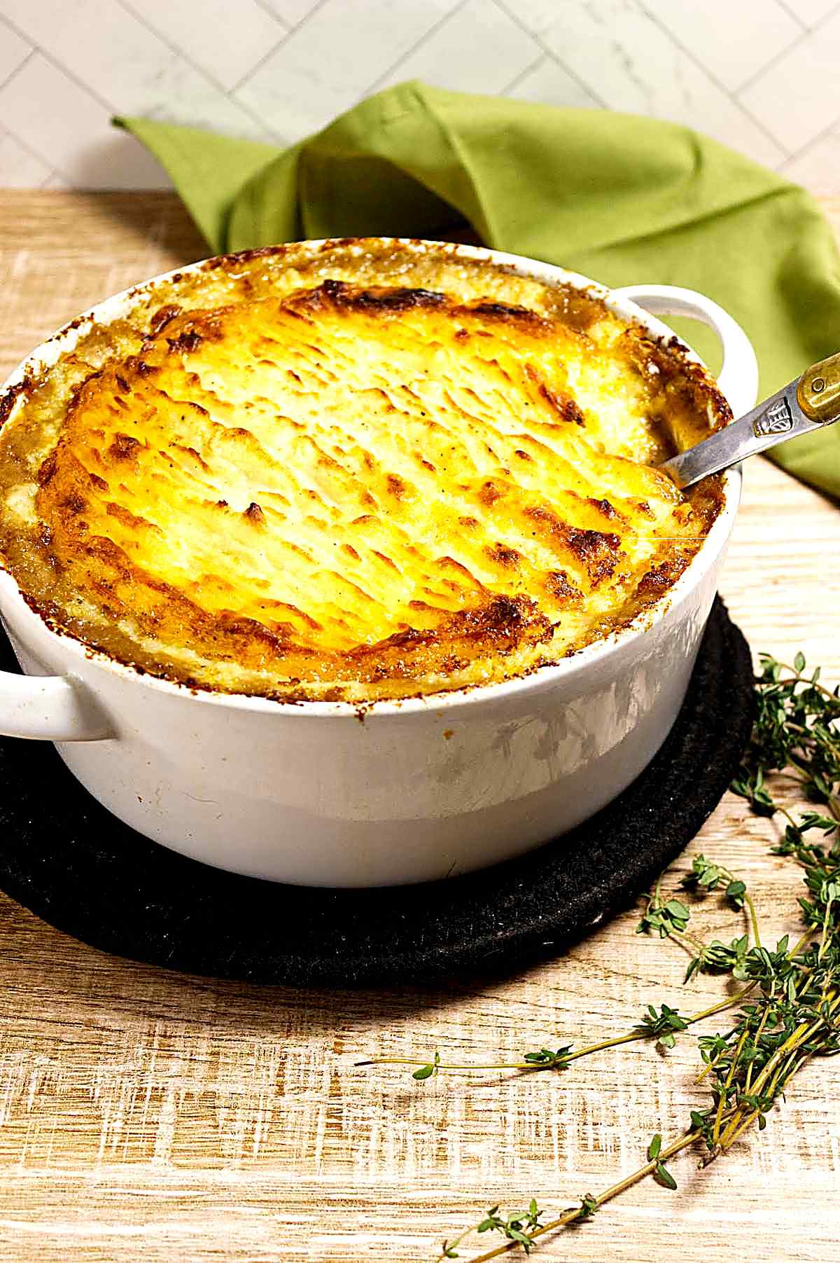 A golden-brown traditional shepherd’s pie in a white casserole dish, sitting on a dark trivet atop a wooden surface, with fresh thyme sprigs and a green cloth napkin nearby. A spoon rests inside the dish.