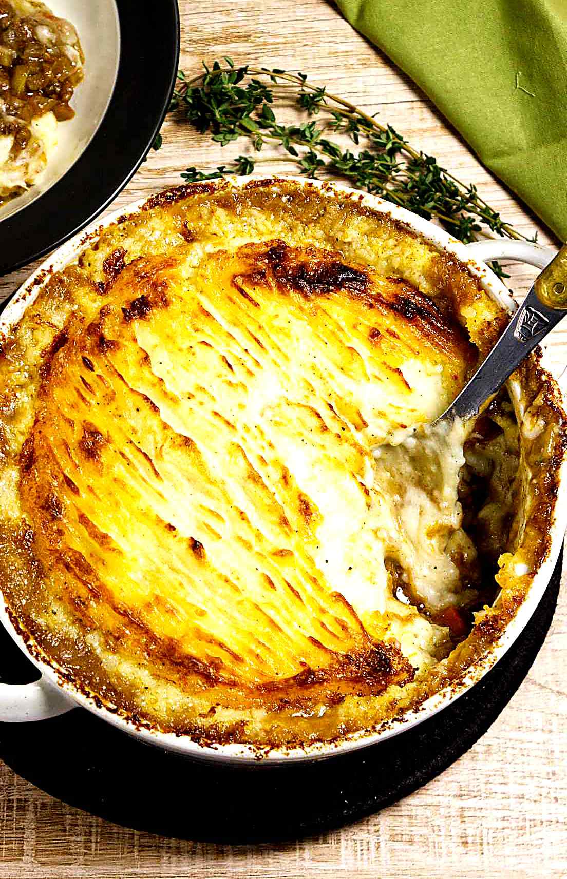 A baked shepherd’s pie with lamb in a round white dish with a golden, ridged mashed potato topping. A serving spoon rests inside, revealing the savory filling. Fresh thyme and a green napkin are nearby on a wooden table.