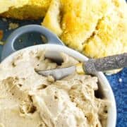 A bowl of creamy cinnamon honey butter with a spreading knife, served next to two pieces of cornbread on a blue plate. Text on the image reads, "Texas Road House Cinnamon Honey Butter.