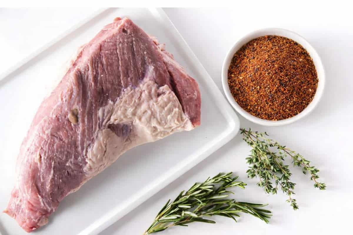 A raw beef tri-tip on a white tray sits next to a bowl of dry spice rub, with fresh rosemary and thyme sprigs arranged beside them on a white surface.