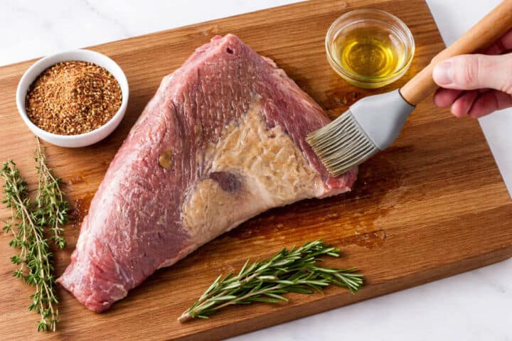 A raw tri-tip steak on a wooden cutting board is being brushed with oil. Fresh rosemary, thyme, a bowl of spice rub, and a small bowl of oil are arranged next to the meat.